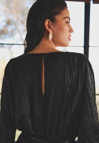 Schwarzes plissiertes Kleid mit einem Ausschnitt am Rücken, weicher Textur und taillierter Passform. Das Model trägt goldene Federohrringe. Natürliches Licht.