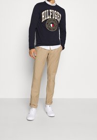 T-paita pitkällä hihalla tummansinisellä värillä, jossa on "HILFIGER"-logo, vaaleat chinos-housut ja valkoiset tennarit. Paidassa on pyöreä kaula-aukko ja painettu emblem.