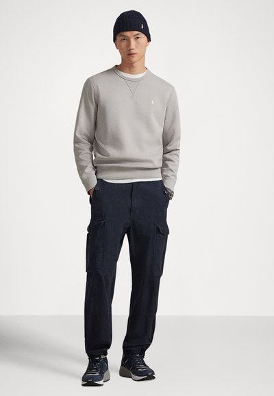 Sweatshirt cinzento claro com decote redondo, combinado com calças cargo azul-marinho escuras com bolsos laterais. Modelo usa um gorro de malha azul-marinho e ténis.