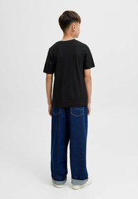 Dječak okrenut leđima nosi crnu majicu kratkih rukava, labave plave traperice podvrnute na rubovima i bijele tenisice na jednostavnoj svijetloj pozadini.