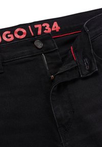 Pantalones vaqueros de mezclilla negra con cremallera oscura y cierre de botón. Presentan un forro interior rojo y un logo rojo contrastante en la cintura.