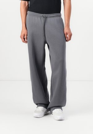 Mand iført løse grå joggingbukser med snøre og hvide sneakers, beskåret ved midten af overkroppen mod en ensartet hvid baggrund.