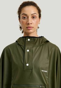 Vihreä vedenpitävä takki hupulla, jossa on korkea kaulus, nepparit, etutasku ja valkoinen logo-ruudutus. Sileä rakenne.