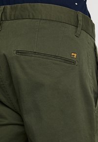 Olivgrüne Hose mit einem Reißverschluss-Rückentasche und einem kleinen goldenen Logoakzent. Hergestellt aus glattem Stoff mit einem taillierten Design.