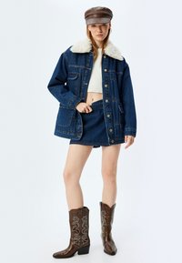 Dżinsowa kurtka z puszystym białym kołnierzem, w zestawie z pasującą spódnicą dżinsową. Brązowe kowbojskie buty mają skomplikowane białe przeszycia.