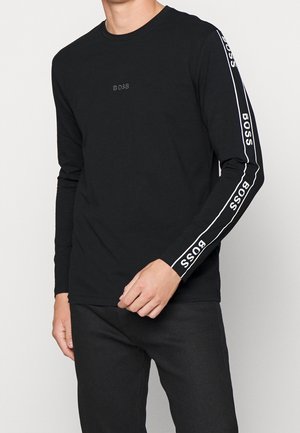 Person trägt ein schwarzes Langarmshirt mit dem "BOSS"-Logo auf der Brust und wiederholtem "BOSS"-Text entlang beider Ärmel in weißen Streifen.
