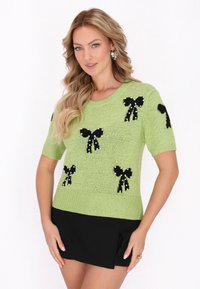 Lysegrøn strikket sweater med sorte sløjfedesigns og perler. Korte ærmer og en tætsiddende silhuet, parret med en sort nederdel.