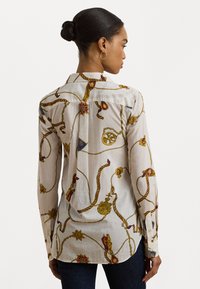 Camicia a maniche lunghe con bottoni in tessuto leggero, con una base bianca, caratterizzata da intricati motivi dorati e colorati, che mostra una texture liscia.