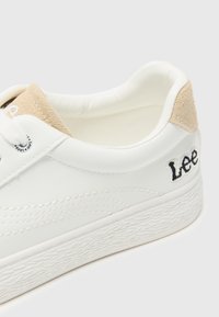 White leather sneaker with suede tan accents at the heel. Features tonal stitching and embossed logo on the side. Textured rubber sole.