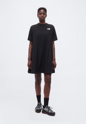 Άτομο στέκεται φορώντας ένα άνετο μαύρο φόρεμα τύπου μπλουζάκι με το λογότυπο The North Face, μαύρες κάλτσες και μαύρα και γκρι αθλητικά παπούτσια με κορδόνια.