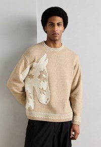 Άνδρας που φοράει μπεζ πουλόβερ με ανάγλυφο σχέδιο κεφαλής άσπρου αλόγου και αστέρια, συνδυασμένο με μαύρο παντελόνι, στέκεται μπροστά σε ανοιχτογκρι τοίχο.