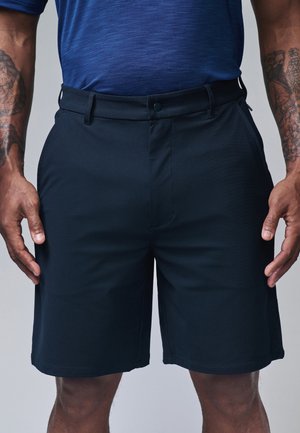 Mężczyzna w granatowych spodenkach i niebieskiej teksturowanej koszuli, stoi z luźno opuszczonymi ramionami wzdłuż ciała, widoczne tatuaże na obu przedramionach.