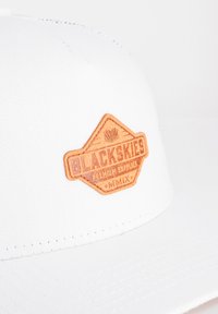 Weißes Baseballcap mit einem strukturierten, tanfarbenen Lederschild, auf dem "BLACKSKIES PREMIUM SUPPLIES MMIX" in Dunkelbraun eingeprägt ist.