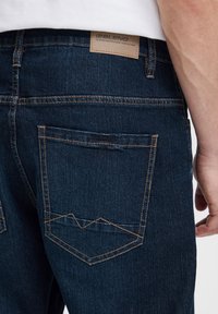 Tummansiniset farkut, joissa on vyötäröllä vaaleanruskea nahkainen merkki ja kontrastina oranssit tikkauskohdat. Takataskussa on hillitty siksak-muotoilu.