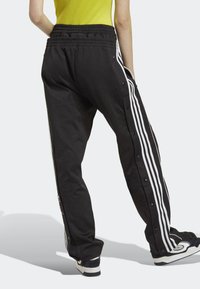 Svarta träningsbyxor med vita sidostripes, elastisk midja och tryckknappar längs sidorna. Mjuk textur med en avslappnad passform.