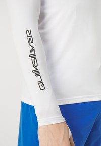 Baltas garās piedurknes krekls ar melnu "Quiksilver" logotipu uz piedurknes, saskaņots ar zilām šortiem, kas izgatavoti no gludas, elastīgas auduma.