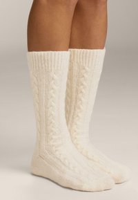 Nogavice do kolen v krem barvi z vzorcem pletenja v obliki kablov in rebrastimi manšetami, izdelane iz mehke, teksturirane tkanine.