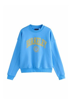 Young woman with light brown hair stands on a street wearing a bright blue sweatshirt and black pants, looking to the side.