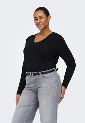 Vrouw die glimlacht, draagt een zwarte top met lange mouwen, grijze spijkerbroek, zwarte riem met dubbele gespsluiting en zilverkleurige oorringen, poserend tegen een effen achtergrond.