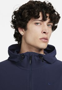 Marinblå hujacka med dragkedja framtill, justerbara dragskoar och en logotyp på bröstet. Har en mjuk tygstruktur och en strutturerad huva.