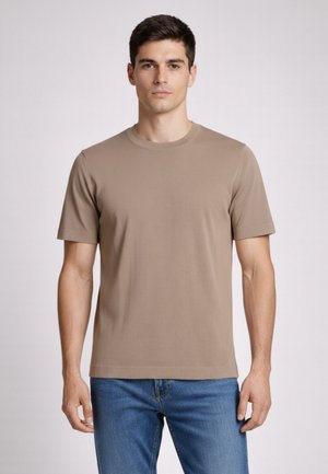 Giovane uomo che indossa una T-shirt beige semplice a maniche corte e jeans blu, in piedi davanti a uno sfondo grigio chiaro.