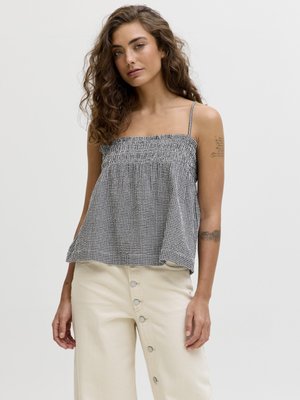 Jeune femme aux cheveux bouclés portant un haut sans manches à carreaux noir et blanc et un pantalon crème avec des boutons latéraux, debout devant un fond uni.