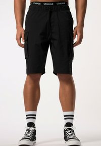 Pantalones cortos de carga negros con bolsillos laterales, cintura elástica y cordón. Combinados con zapatillas negras de caña alta y calcetines blancos a rayas.