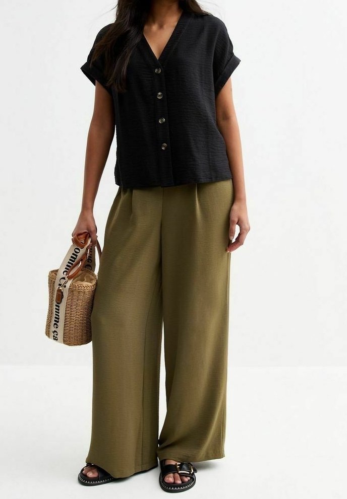 Femme portant un chemisier noir à manches courtes boutonné, un pantalon large vert olive, des sandales noires, et tenant un sac tissé avec une bandoulière imprimée.