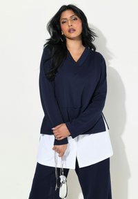 Camiseta de manga larga en azul marino con escote en V, que presenta un diseño estructurado y aberturas laterales. El modelo sostiene un pequeño bolso plateado, superpuesto sobre una camisa blanca.