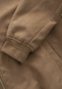 Manche de veste marron avec bord-côte et fermeture éclair visible le long de la couture latérale sur un fond de tissu doux.
