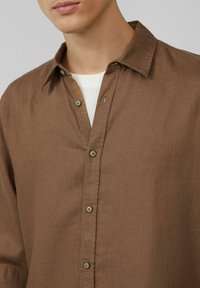 Chemise brune à boutons en tissu texturé, dotée d'un col, d'une patte de boutonnage avec des boutons en bois, et portée par-dessus une chemise blanche.