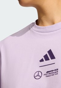 Svijetlo lavanda majica s okruglim izrezom, s tamno ljubičastim Adidas logotipom i Mercedes-Benz oznakom otisnutim na prednjoj strani. Mekana tekstura tkanine.