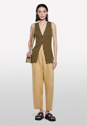 Gilet sans manches à côtes vert olive avec boutons à l'avant, associé à un pantalon large jaune clair et des sandales noires avec un détail de sangle.