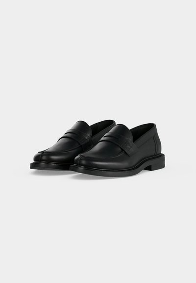 Paire de mocassins noirs en cuir lisse, sans lacets, avec petits talons et une simple bride sur le dessus, sur un fond clair et uni.
