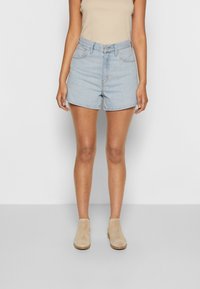 Shorts de mezclilla azul claro con cintura alta, bolsillos delanteros y un ajuste holgado, combinados con botas hasta el tobillo beige y una blusa ajustada beige.