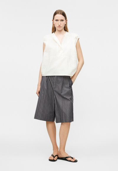 Γυναίκα που φορά λευκό αμάνικο μπλουζάκι, μαύρο και λευκό παντελόνι με κάθετες ρίγες και φαρδιές πόδες, και μαύρες σαγιονάρες, όρθια με το ένα χέρι στην τσέπη.