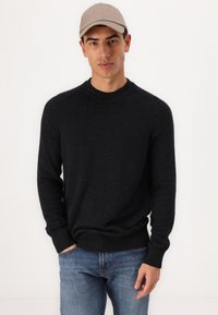 Svart strikket genser med rund hals og ribbestrikkede mansjetter og kant, båret over blå jeans, kombinert med en lys beige caps.