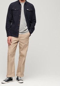 Camicia blu scuro con bottoni sopra una maglietta grigia, pantaloni beige e sneakers nere. Caratteristiche includono due tasche sul petto e maniche arrotolate.