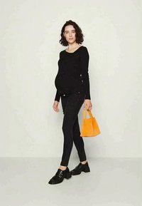 Top negru cu mâneci lungi, colanți negri, încălțăminte neagră cu catarame și o mică geantă portocalie. Modelul stă pe un fundal neutru.