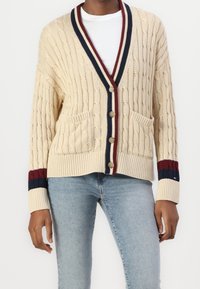 Cardigan beige en maille torsadée avec bords côtelés, rayures bleu marine et grenat, col en V, fermeture par boutons, et deux poches avant. Texture douce.