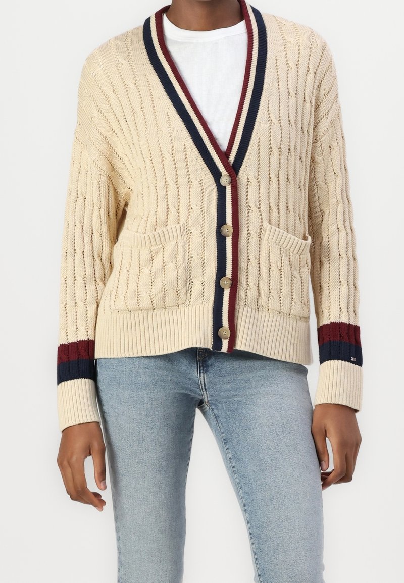 Cardigan beige en maille torsadée avec bords côtelés, rayures bleu marine et grenat, col en V, fermeture par boutons, et deux poches avant. Texture douce.