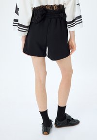 Pantaloni corti neri con vita elastica, texture liscia e vestibilità comoda, abbinati a calzini neri e scarpe. Dettagli visibili delle gambe.