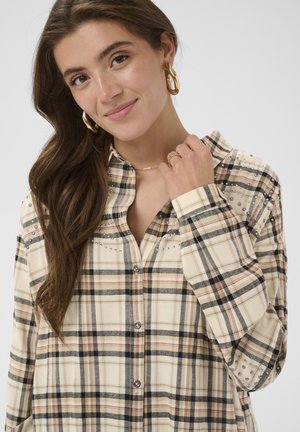Jeune femme aux longs cheveux bruns portant de grandes boucles d'oreilles dorées en forme d'anneau et une chemise à carreaux beige ornée de perles, souriant doucement à la caméra.