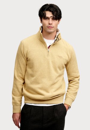 Ung mand med mørkt, bølget hår iført en beige kvartlynlås sweater, hvid undertrøje og mørke bukser, med den ene hånd i lommen.