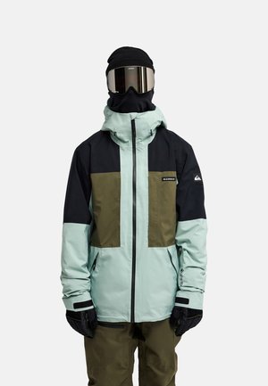 Personne portant un bonnet noir, des lunettes réfléchissantes, une veste d’hiver bleu clair et vert olive, des gants noirs et un pantalon vert olive, debout sur fond blanc.