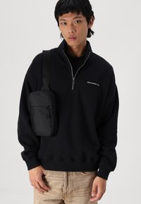 Sweat-shirt noir zippé avec un col haut et un logo brodé, associé à un sac bandoulière compact noir avec des compartiments zippés.