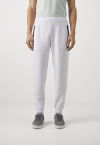 White sweatpants with elastic waistband, two side pockets featuring black zippers, and ribbed cuffs. Worn with gray suede sneakers.