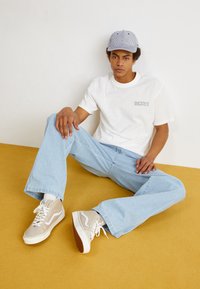 Mladý muž v pruhované čepici, bílé tričko Dickies, světle modré džíny a béžové vysoké tenisky sedící na hořčicově žluté podlaze proti bílé zdi.