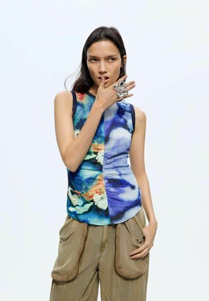 Kobieta ubrana w niebieską i wielokolorową bluzkę bez rękawów oraz luźne brązowe spodnie, dotykająca ust dużym ozdobnym srebrnym pierścieniem.