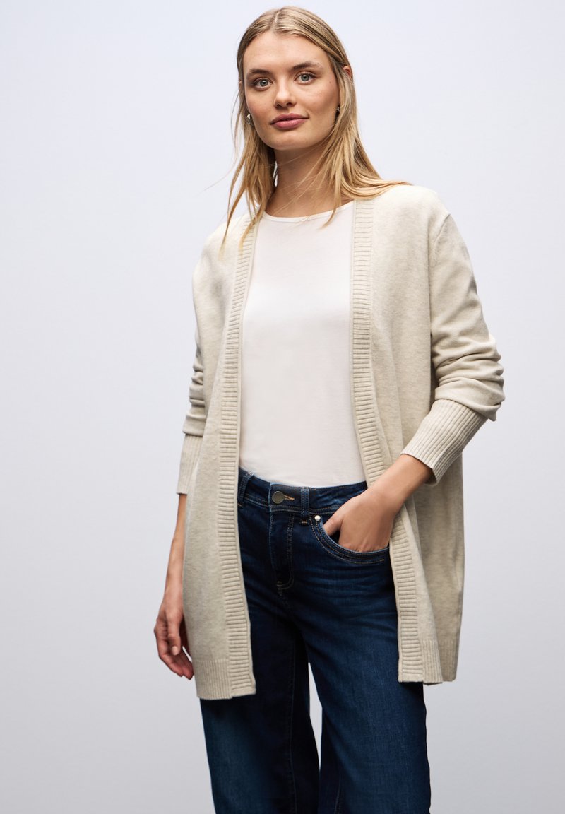 Ljusbeige cardigan i mjukt tyg, med öppen front och ribbade kanter, bärs över en vit topp och tillsammans med mörka jeans.